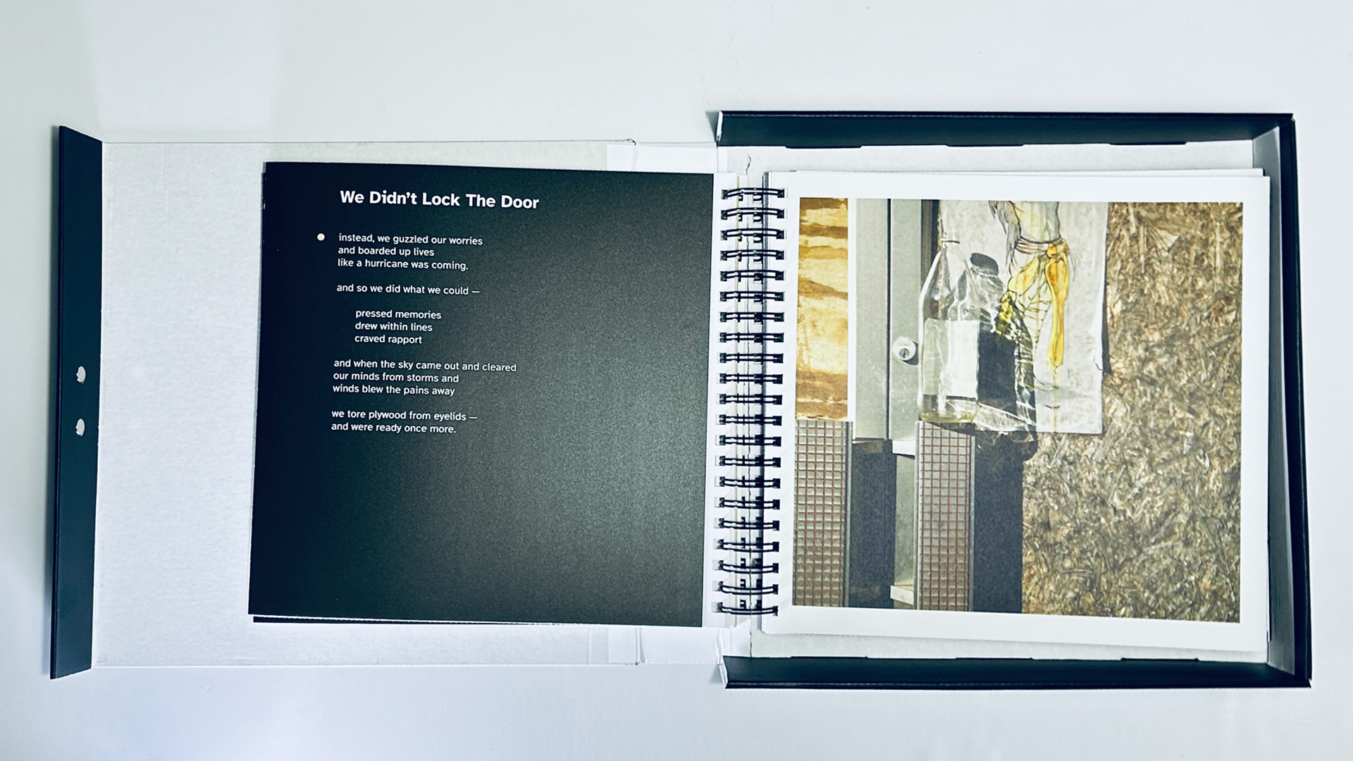 Book_Open_to_a_Spread002 The same handmade, spiral-bound artist book is open to a different two-page spread. On the left page, a poem titled “We Didn’t Lock The Door” is printed in white sans-serif type on matte dark down paper. A bullet point introduces the first stanza, and the text flows in a centered, vertical layout with poetic spacing and line breaks. On the right page is a mixed-media photograph featuring a close-up of a door partially boarded with chipboard. Transparent tape, a clear glass bottle, and a faint pencil-and-watercolor sketch of a yellow garment overlap the surface, creating layers of meaning and material. The textures—gritty wood, soft paper, smooth glass—convey a tactile sense of place and memory. Like the previous spread, the photograph is bordered in white, emphasizing contrast and balance within the spread. The book’s physical structure and design continue to convey intimacy, memory, and materiality.