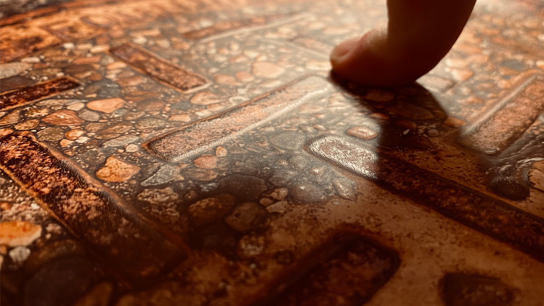 finger_touches_tactile_photo A close-up photograph shows a person's finger gently touching the surface of a tactile print. The image captures a richly textured and embossed photograph of a stone and brick pathway, printed in warm earth tones of burnt orange, copper, and brown. The tactile surface includes raised brick-like shapes embedded within a pebbled texture, inviting both visual and haptic exploration. The lighting is low and directional, casting soft shadows that highlight the dimensionality of the print and the subtle sheen of the raised areas. The skin tone of the finger adds contrast to the warm, rocky surface, emphasizing the interaction between body and material. This intimate moment conveys the sensory intention behind the design — allowing touch to become a primary mode of reading and interpretation.